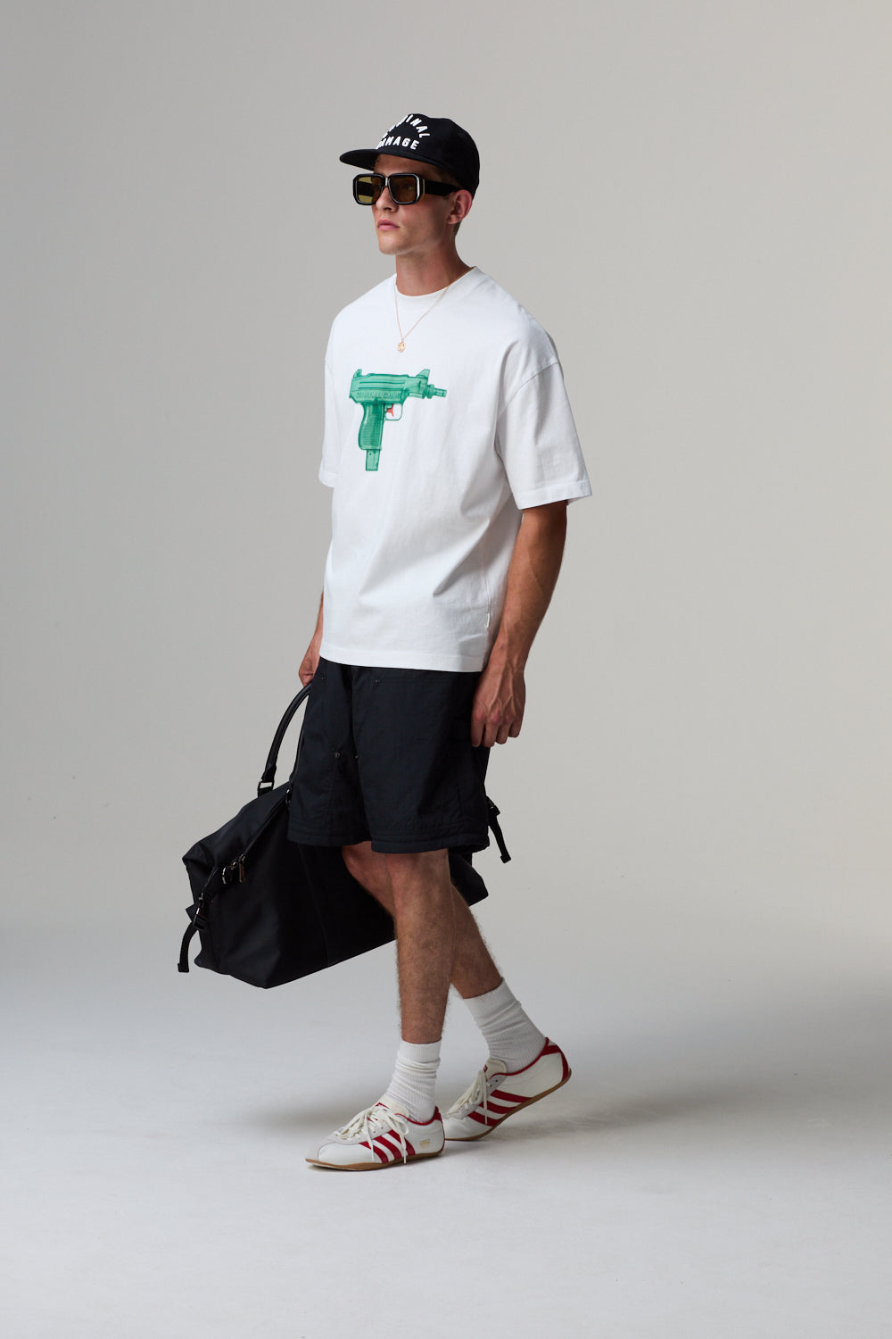 Model wearing a white Criminal Damage t-shirt with a green graphic, black shorts, and a black cap, holding a black bag.