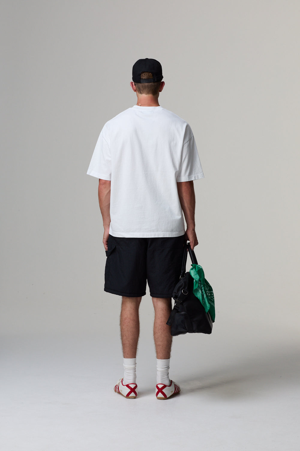 Model wearing a white Criminal Damage t-shirt, black shorts, and a cap, holding a black bag on a grey background