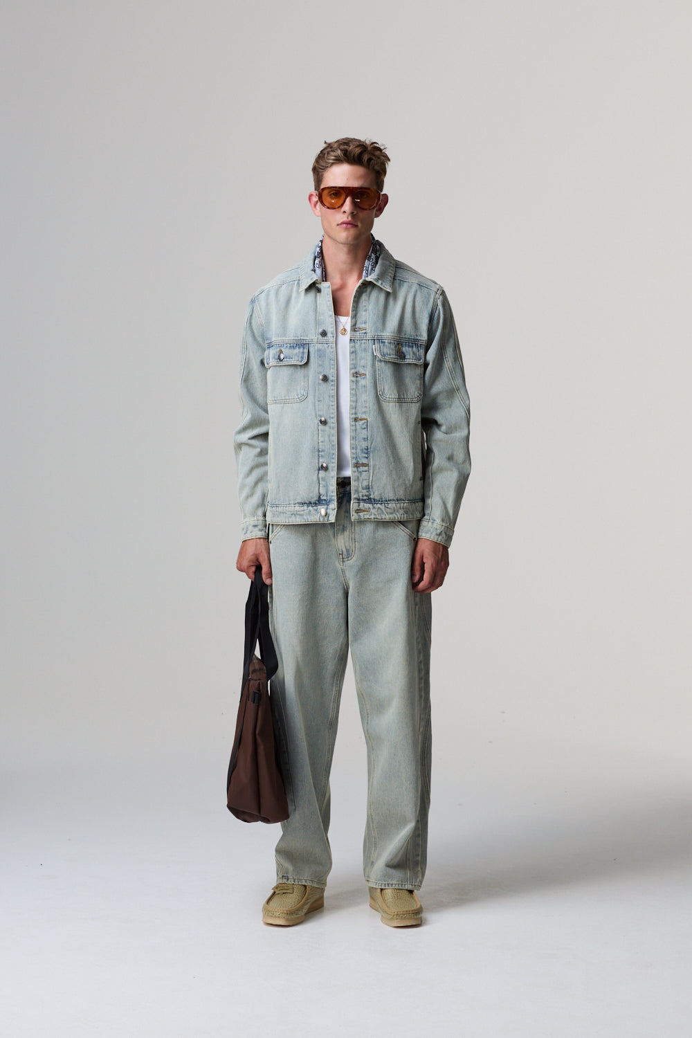 Model wearing a light blue Criminal Damage denim jacket and pants holding a brown leather bag on a grey background