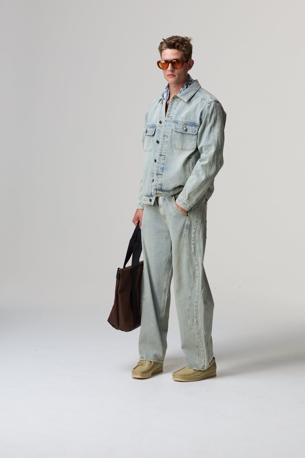 Model wearing a light blue Criminal Damage denim jacket and pants with sunglasses and a brown bag on a grey background