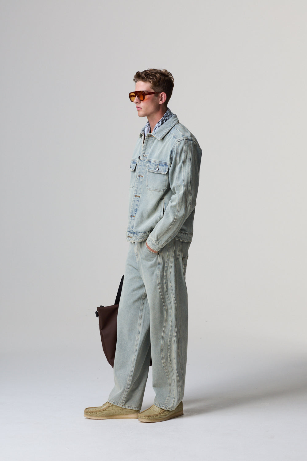 Model wearing a light blue Criminal Damage denim outfit with sunglasses and a bag on a grey background