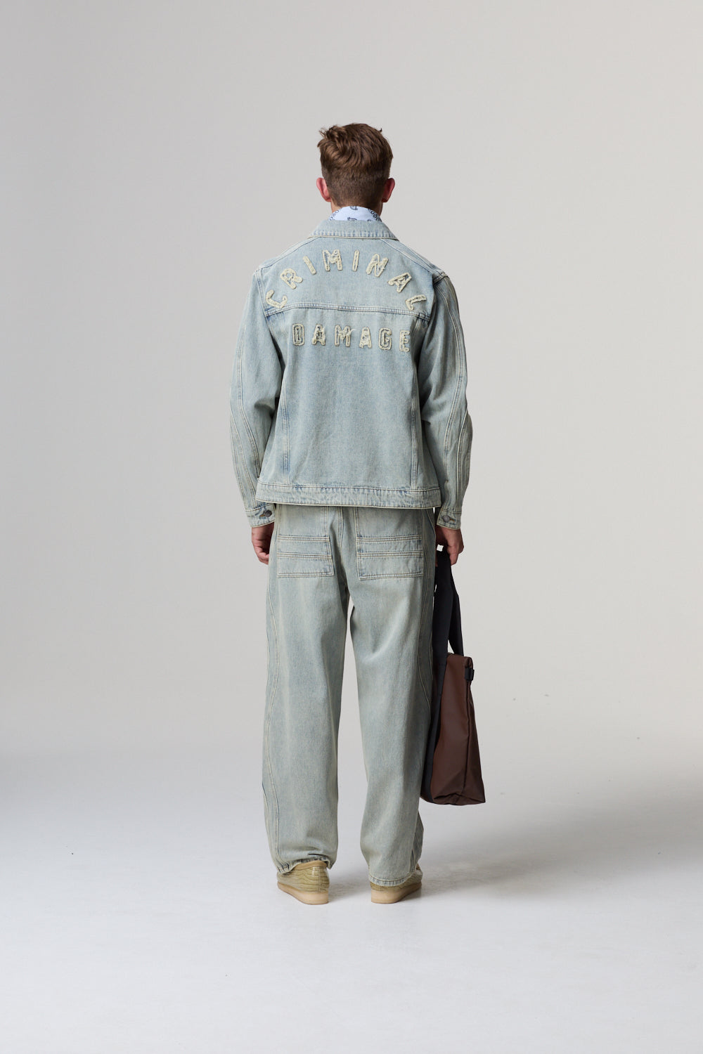 Model wearing a light blue Criminal Damage denim jacket and pants with text on the back, holding a brown bag.