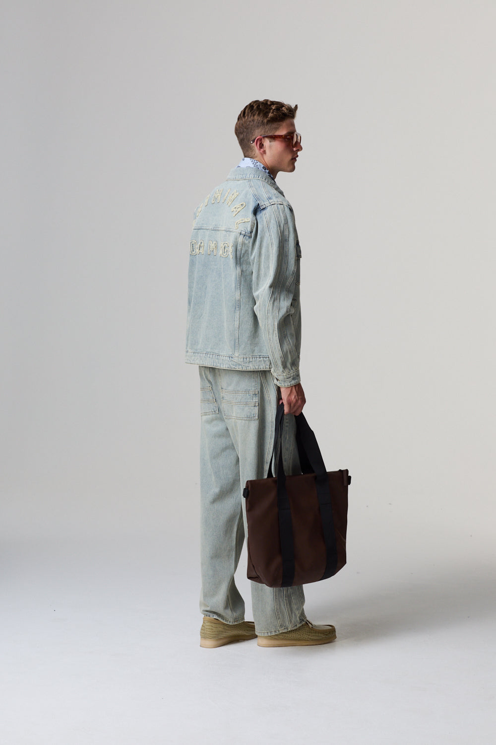 Model holding a brown bag wearing a light blue Criminal Damage denim jacket and pants on a grey background