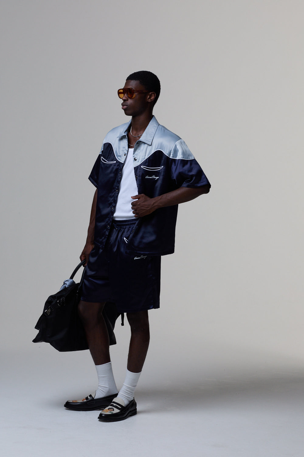 Model wearing a Criminal Damage navy and white jacket with dark shorts, holding a black bag on a grey background