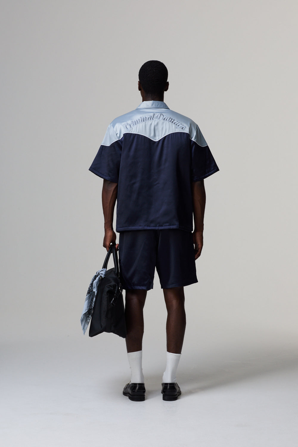 Model wearing a Criminal Damage navy and white shirt with shorts, holding a bag, on a grey background