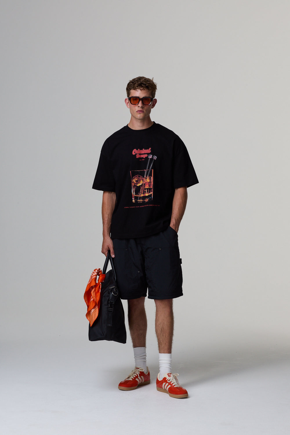 Model wearing a black Criminal Damage graphic t-shirt, black shorts, red sneakers, and holding an orange bag on a grey background