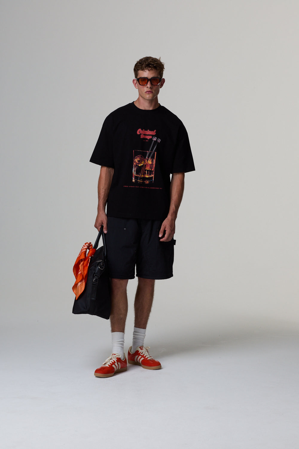 Model wearing a black graphic t-shirt, black shorts, and red shoes on a grey background