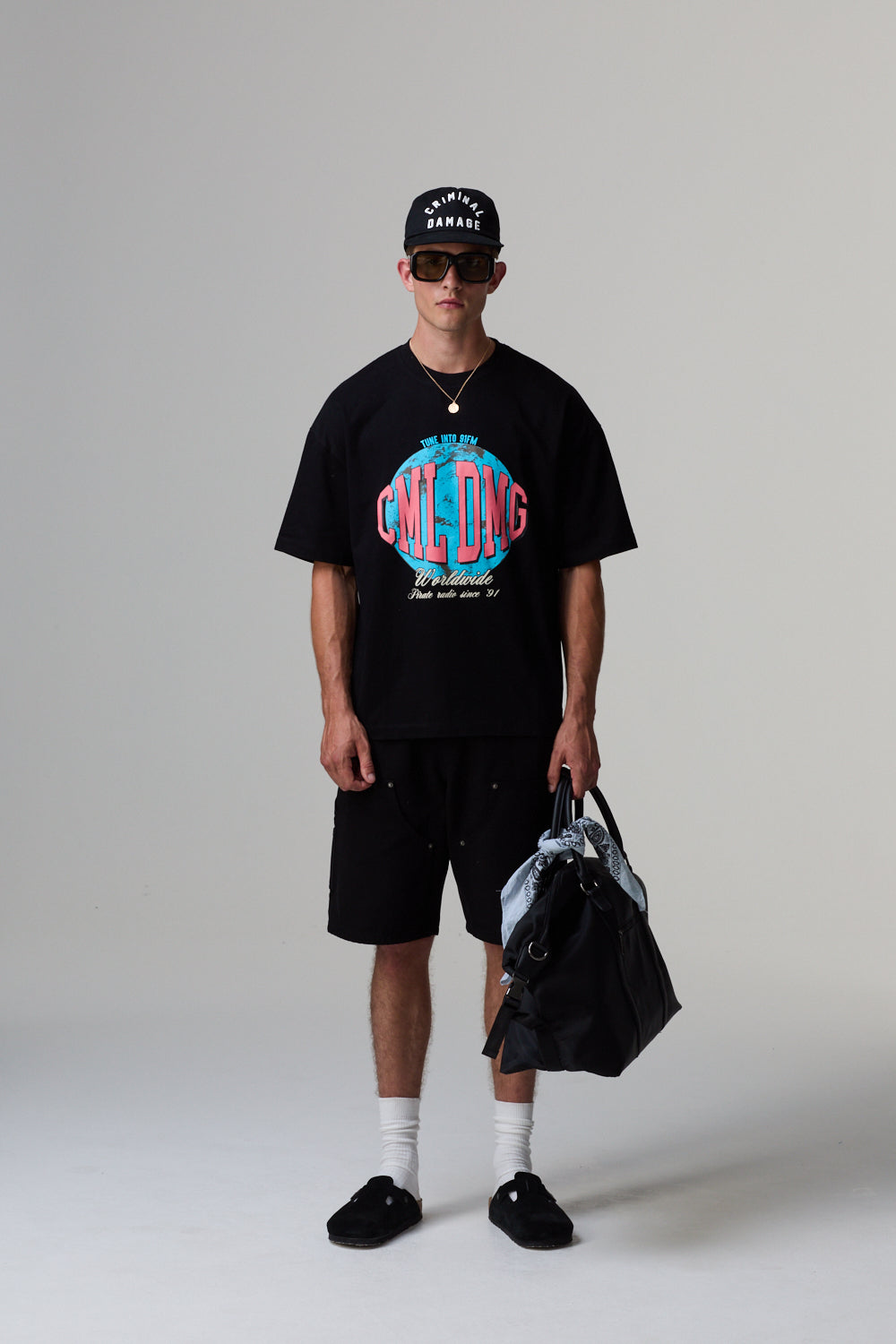 Model wearing a Criminal Damage black t-shirt with colourful graphics, black shorts, and a black cap on a grey background