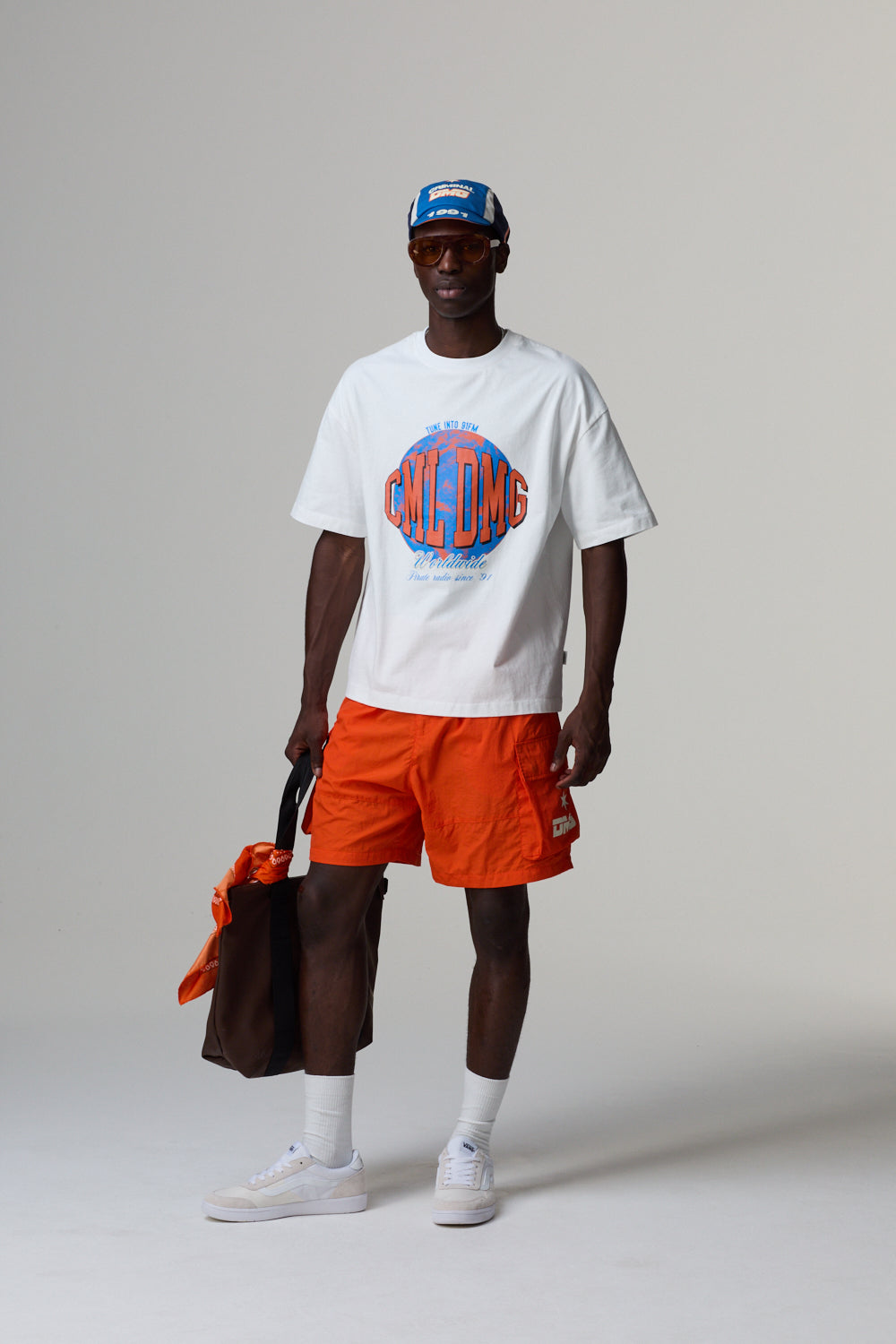 Model wearing a white Criminal Damage t-shirt with a graphic design, orange shorts, and a cap, standing against a grey background.