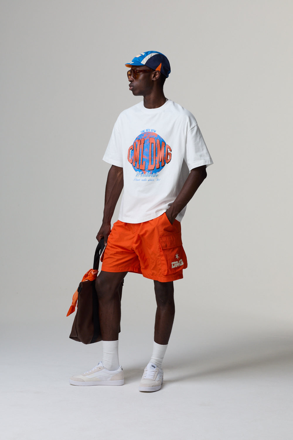 Model wearing a white Criminal Damage t-shirt with a graphic design, orange shorts, and a cap on a grey background