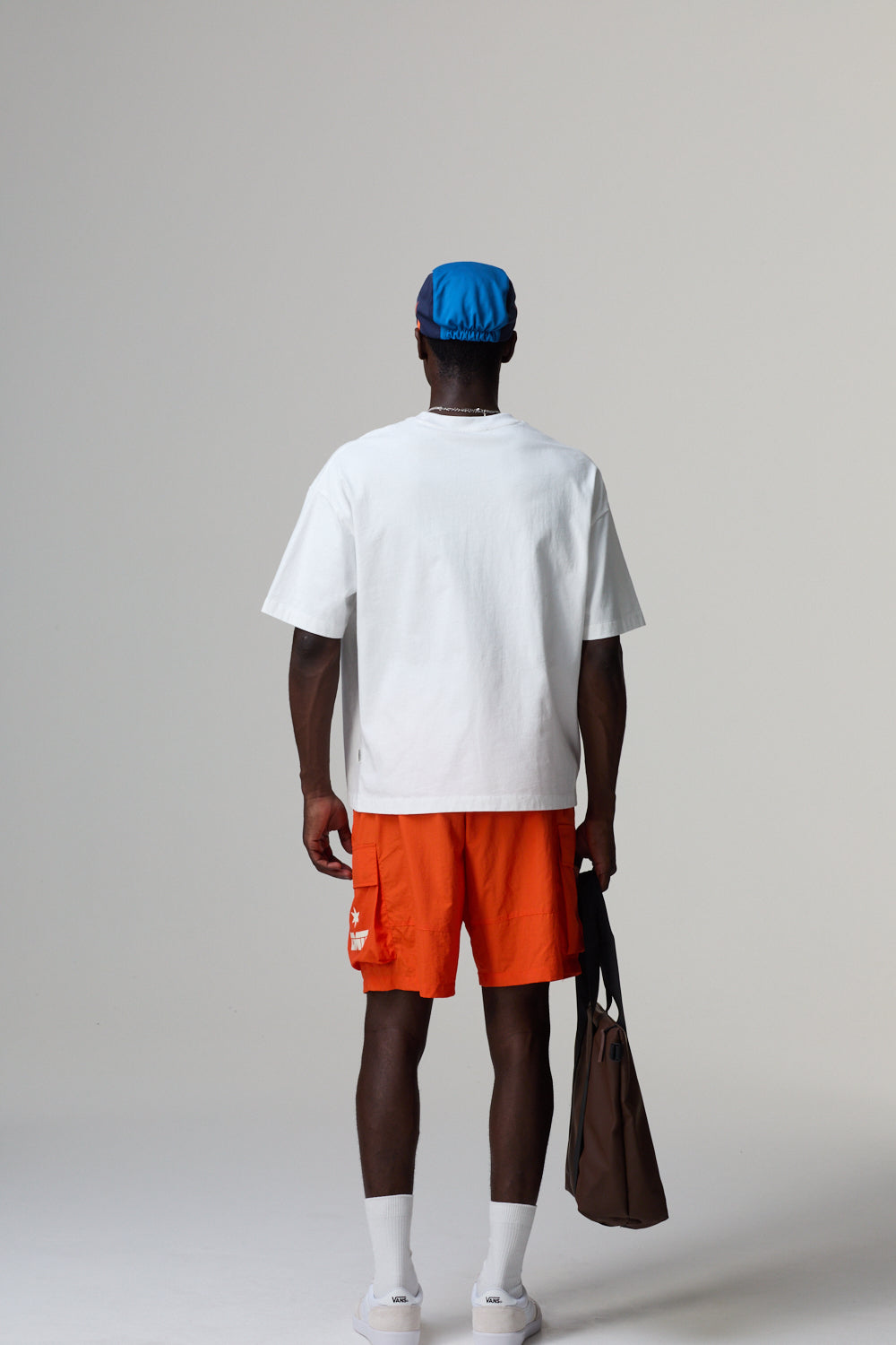 Model wearing a white  criminal Damage t-shirt, orange shorts, and a blue cap on a grey background