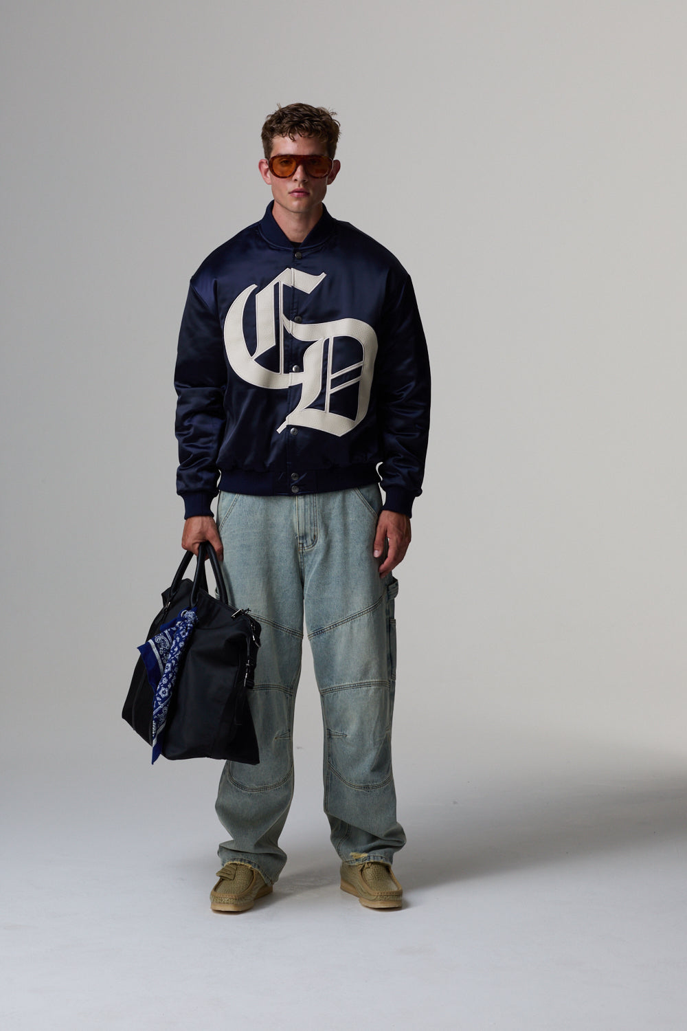 Model wearing a navy Criminal Damage jacket, light blue jeans, and holding a black bag on a grey background