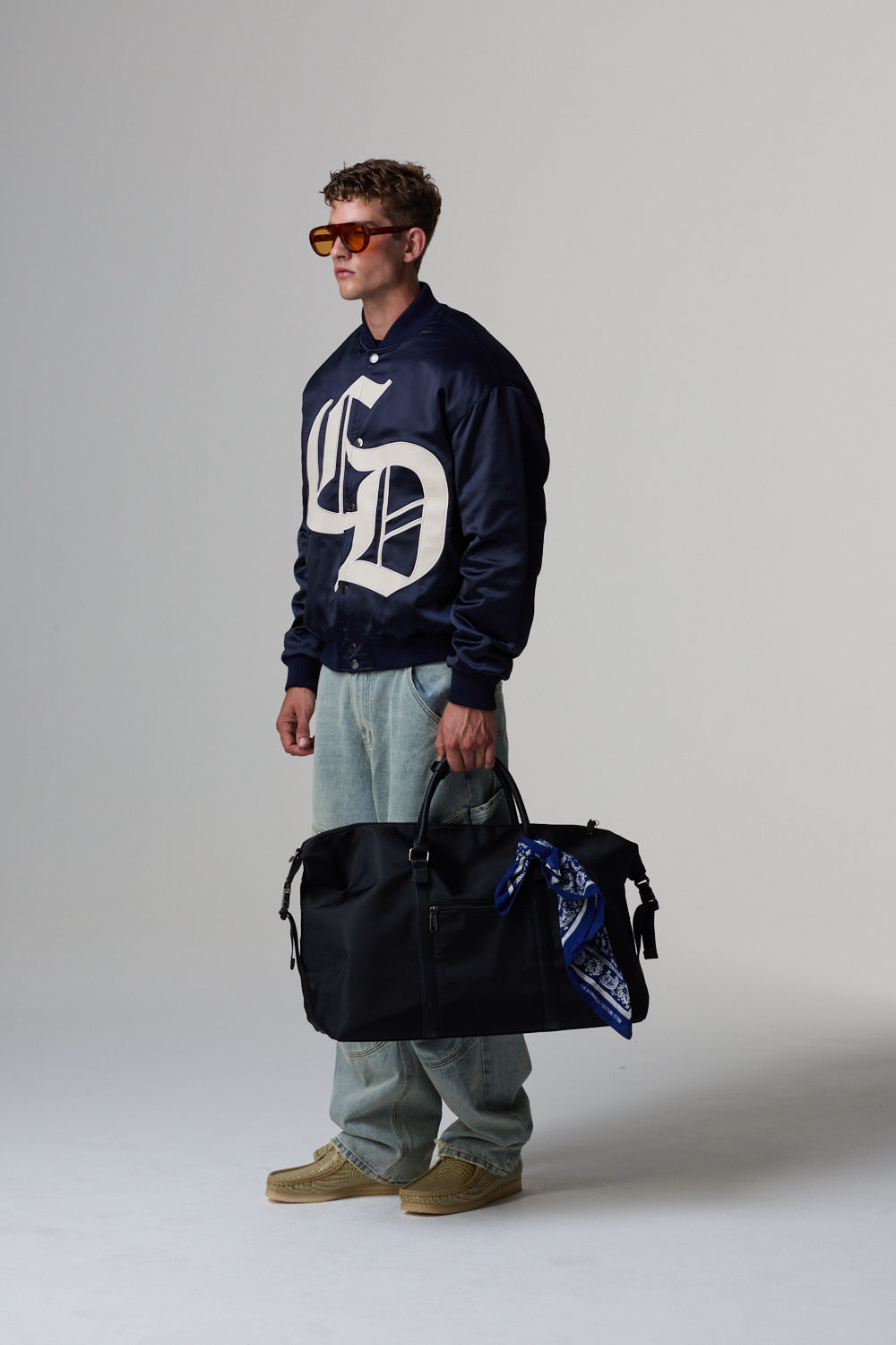 Model wearing a navy Criminal Damage blue jacket, holding a black duffel bag on a grey background