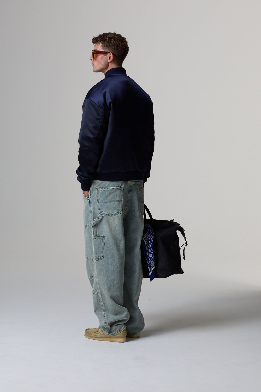 Model in navy Criminal Damage jacket and light blue jeans holding a black bag on a grey background