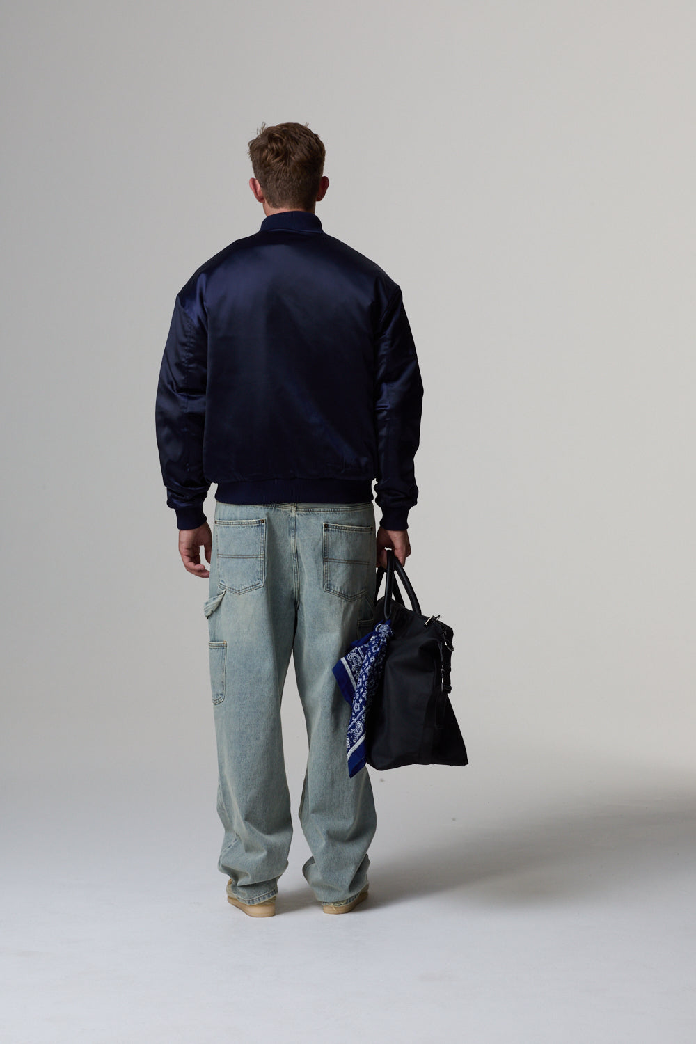 Model wearing a Criminal Damage jacket, holding a black bag and a blue scarf on a grey background